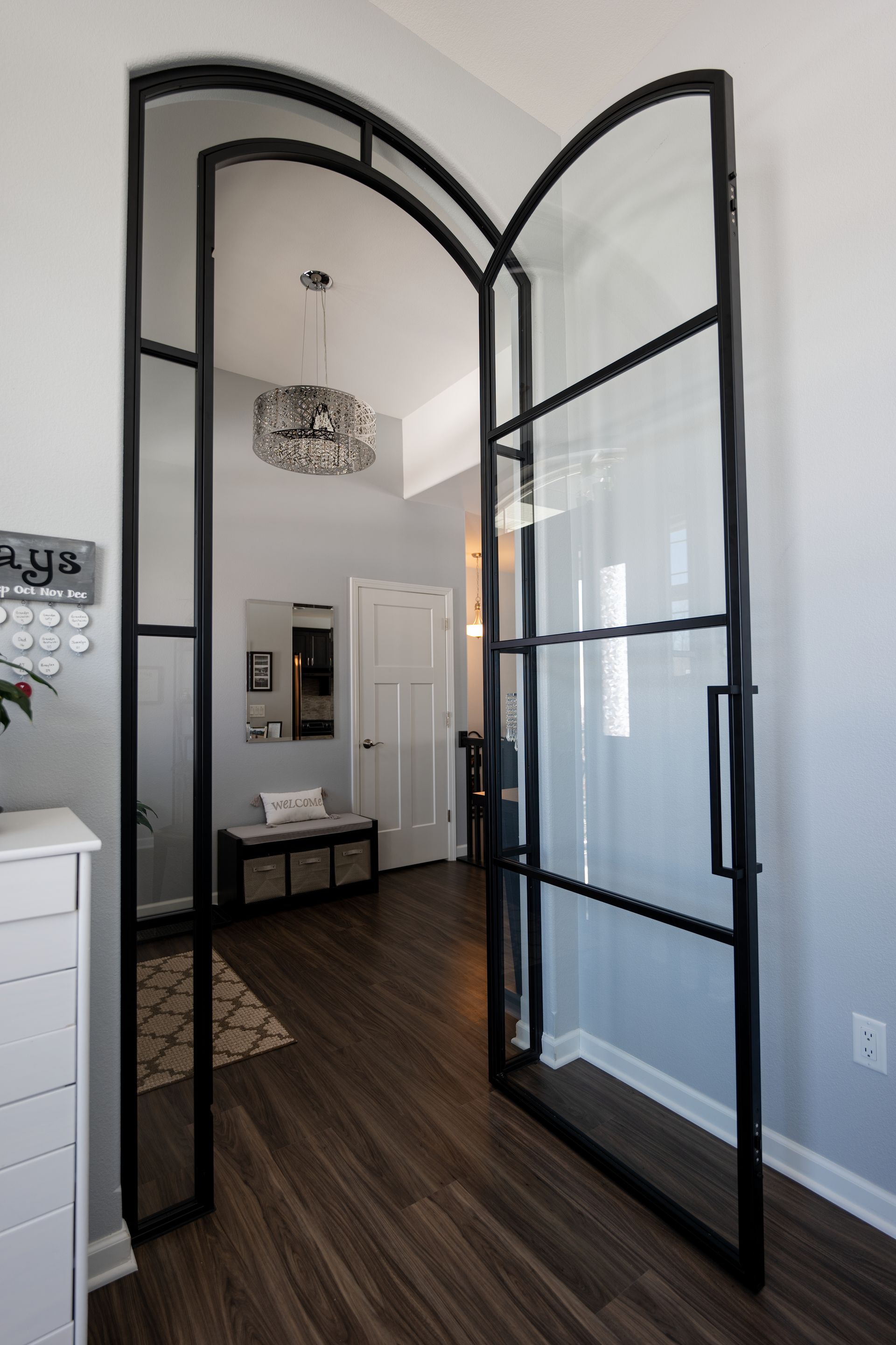 Black-framed arched doorway with clear glass panels, open to a hallway with dark wood floor, white walls and decor.