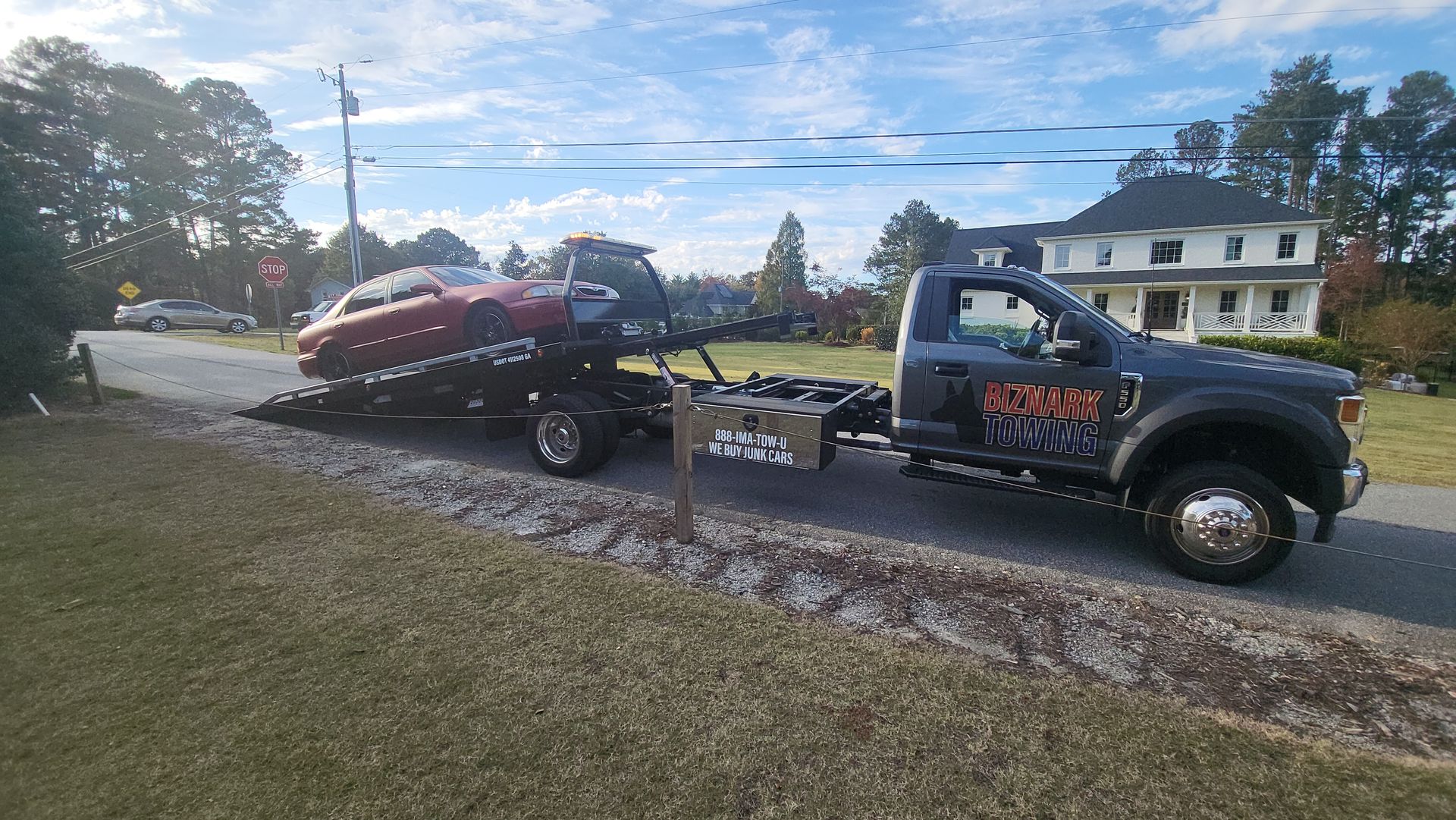 Tow truck carrying a red car, parked on a residential street. A large house is in the background.