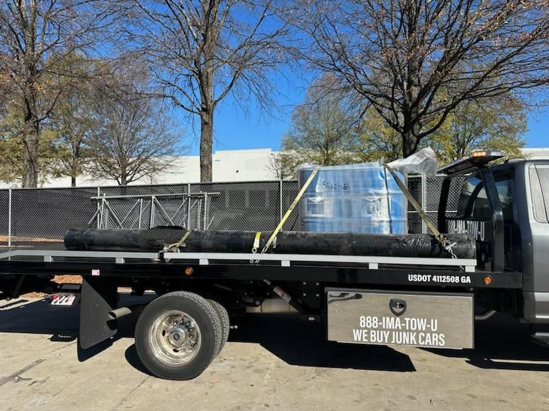 A flatbed tow truck carrying long, black pipes and a wrapped object, outdoors.
