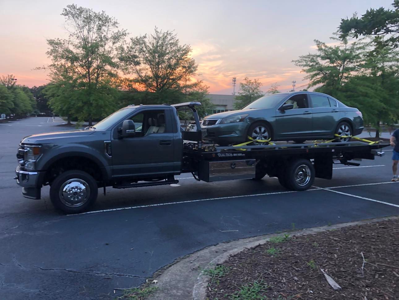 Tow truck carrying a gray car on its flatbed, parked outdoors. Evening light.
