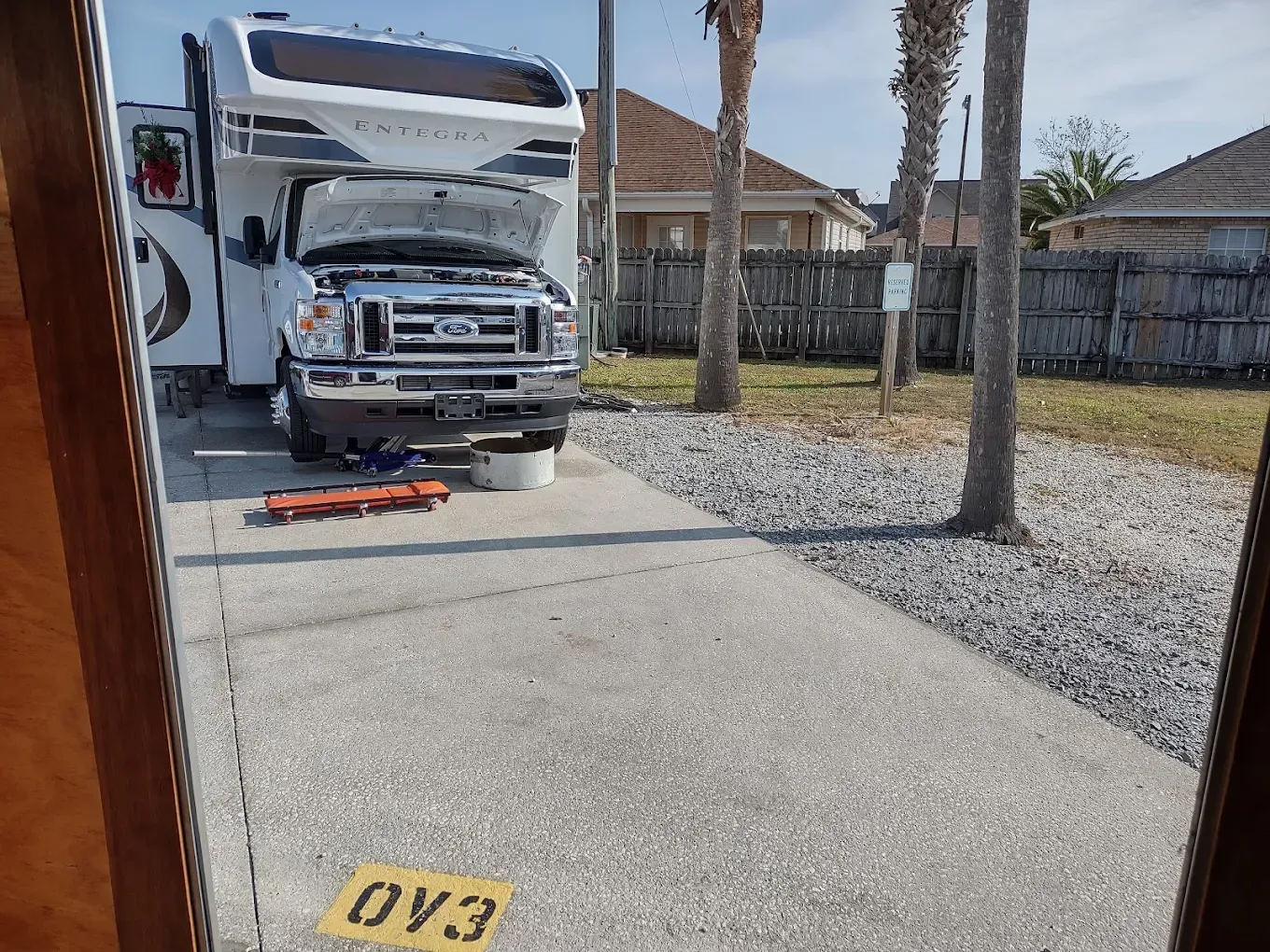RV parked with hood up, tools out. RV site view with palm trees, sunny day.