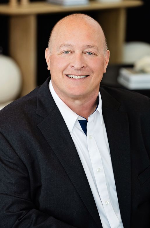 Smiling bald man in a black blazer and white shirt, seated indoors.