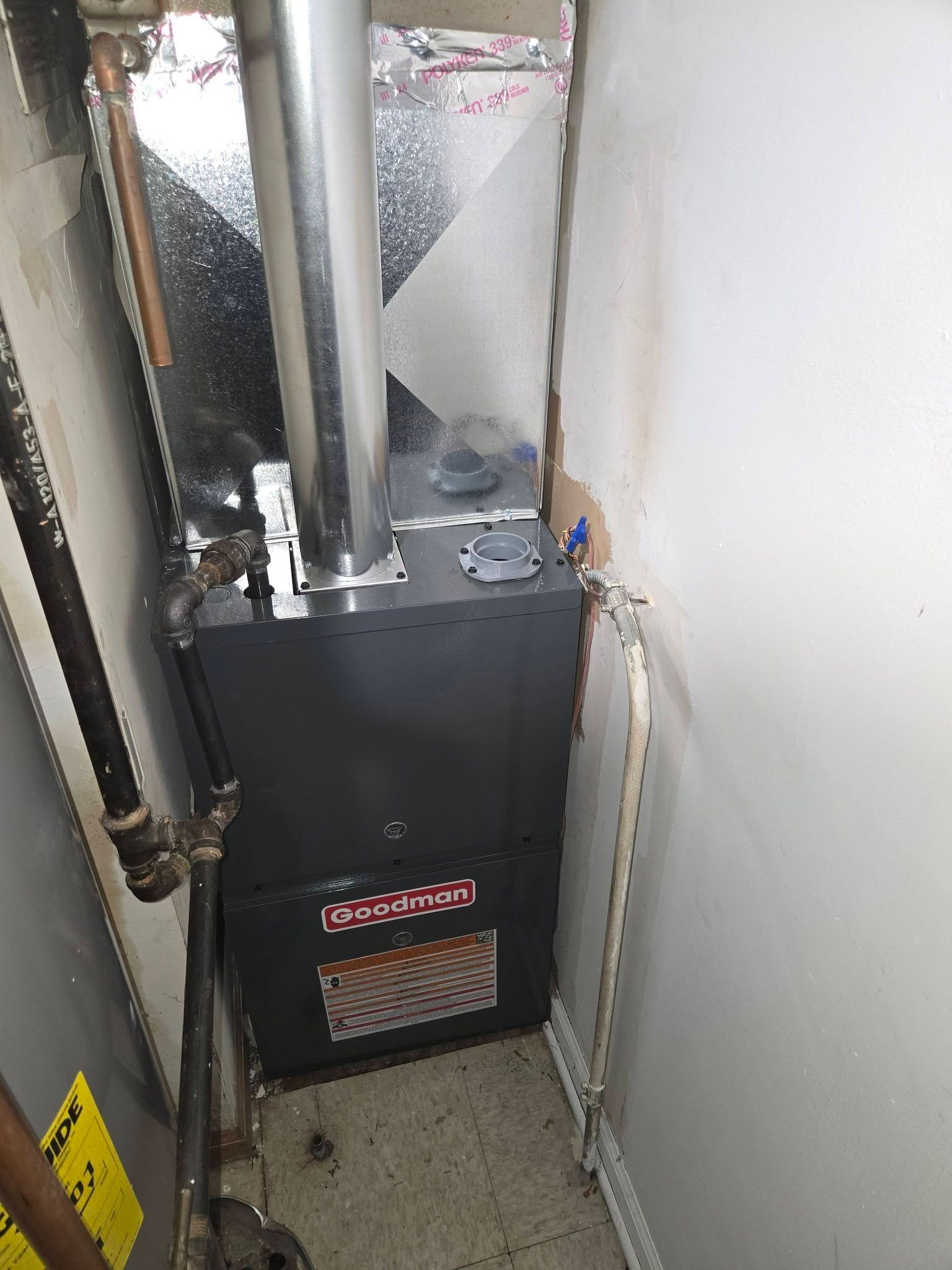 A dark grey Goodman furnace in a basement setting with silver exhaust pipe.