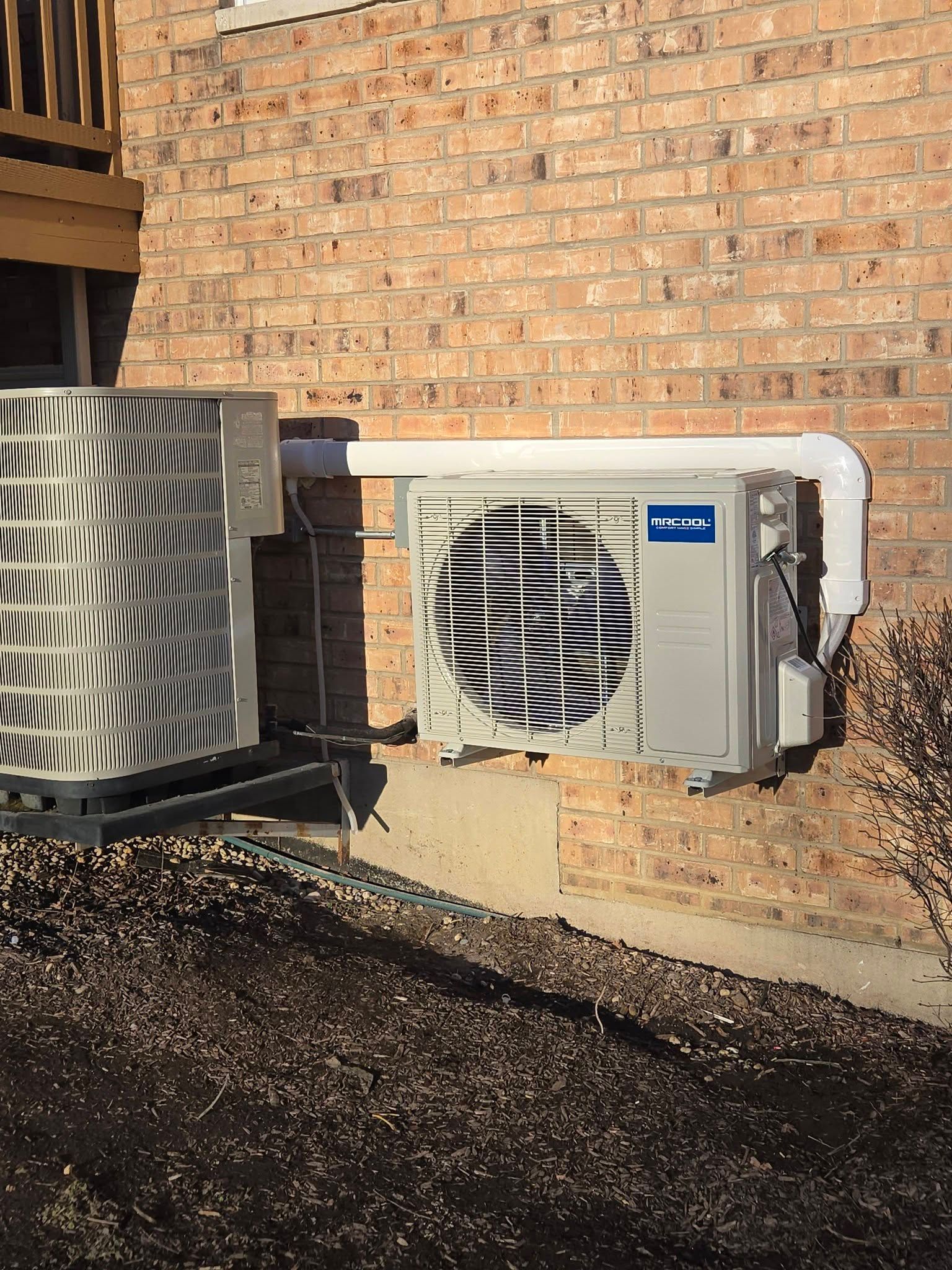 Two air conditioning units mounted on a brick wall, one larger and one smaller with a white pipe connecting them.