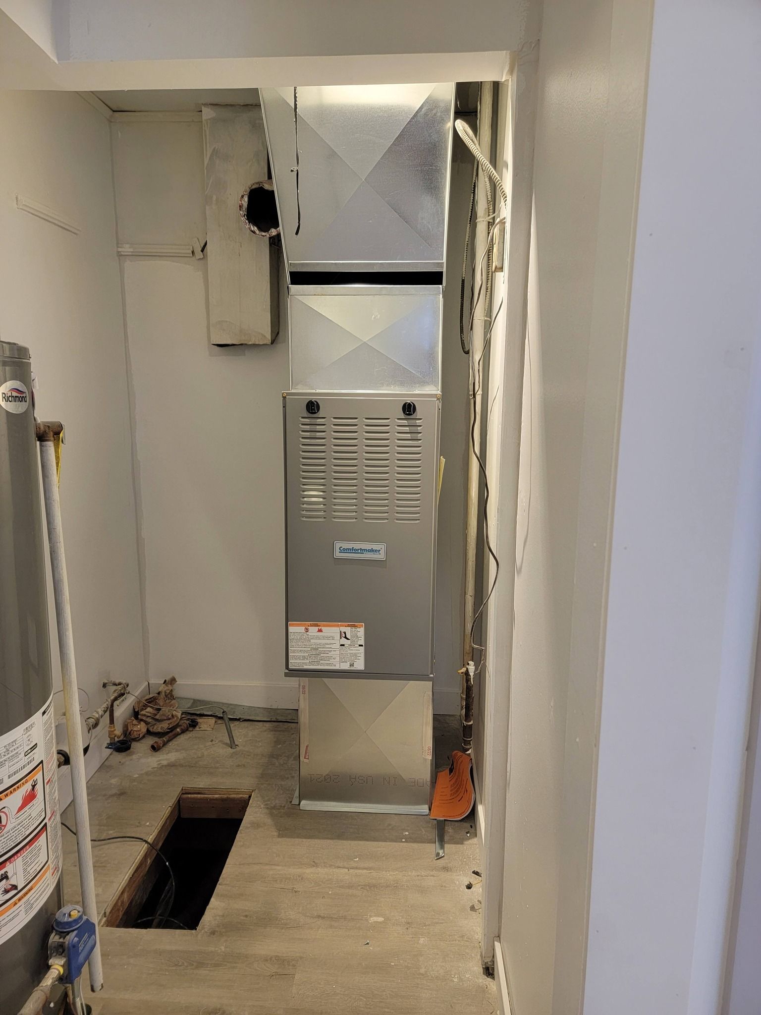 HVAC system in a small room with a water heater and a floor opening. Gray and white interior.