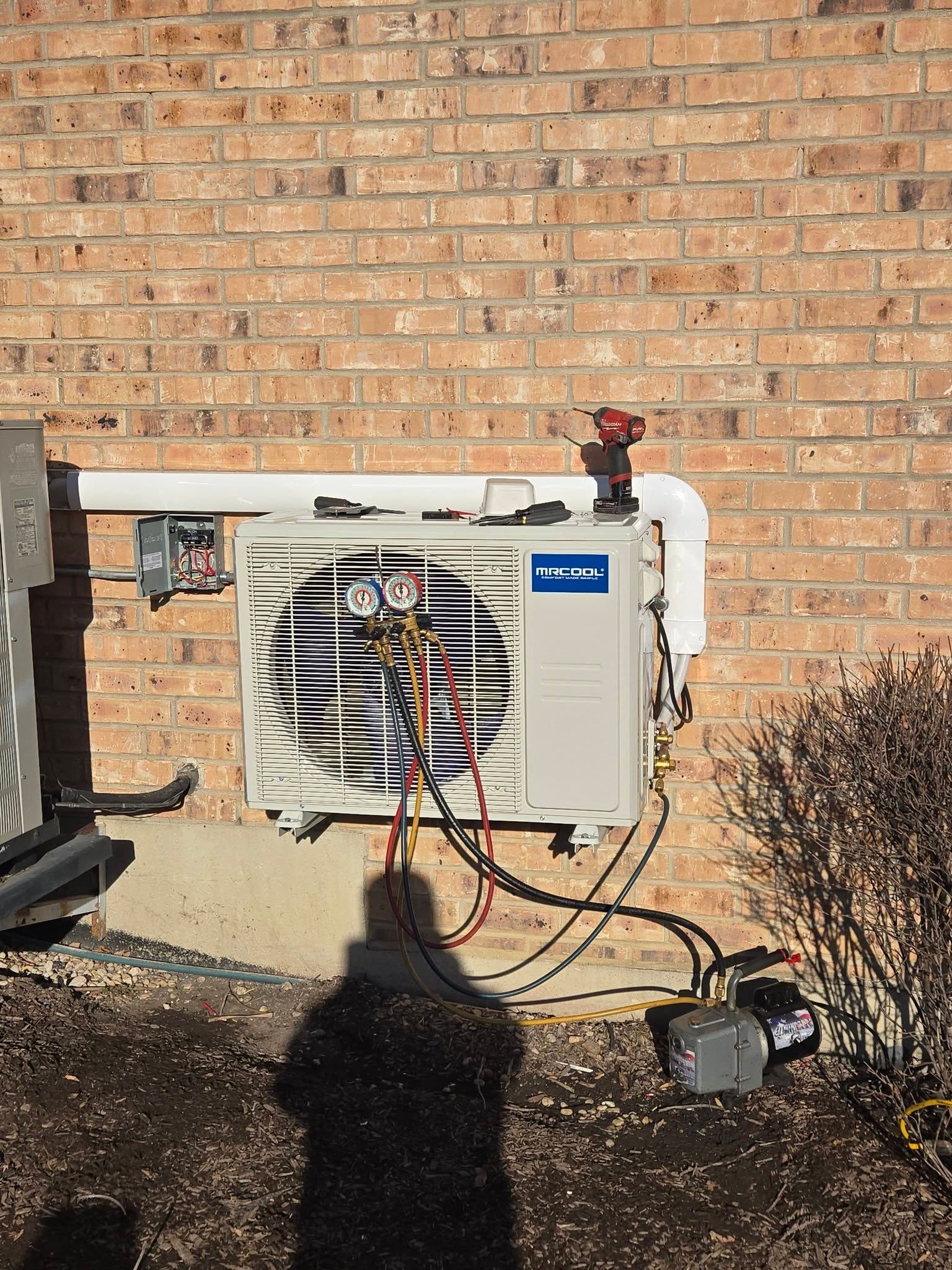 AC unit mounted on a brick wall, with gauges and a vacuum pump connected for servicing.