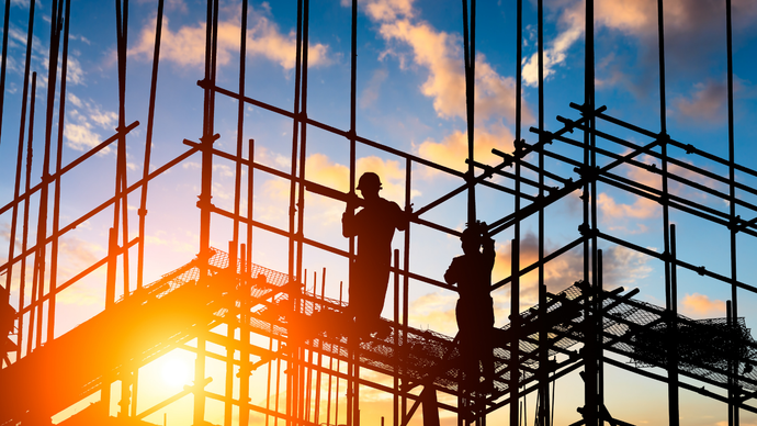 Silhouetted construction workers on scaffolding against a bright sunset sky.