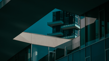 A modern building with teal-toned glass, sharp geometric lines, and a reflection of a high-rise structure.