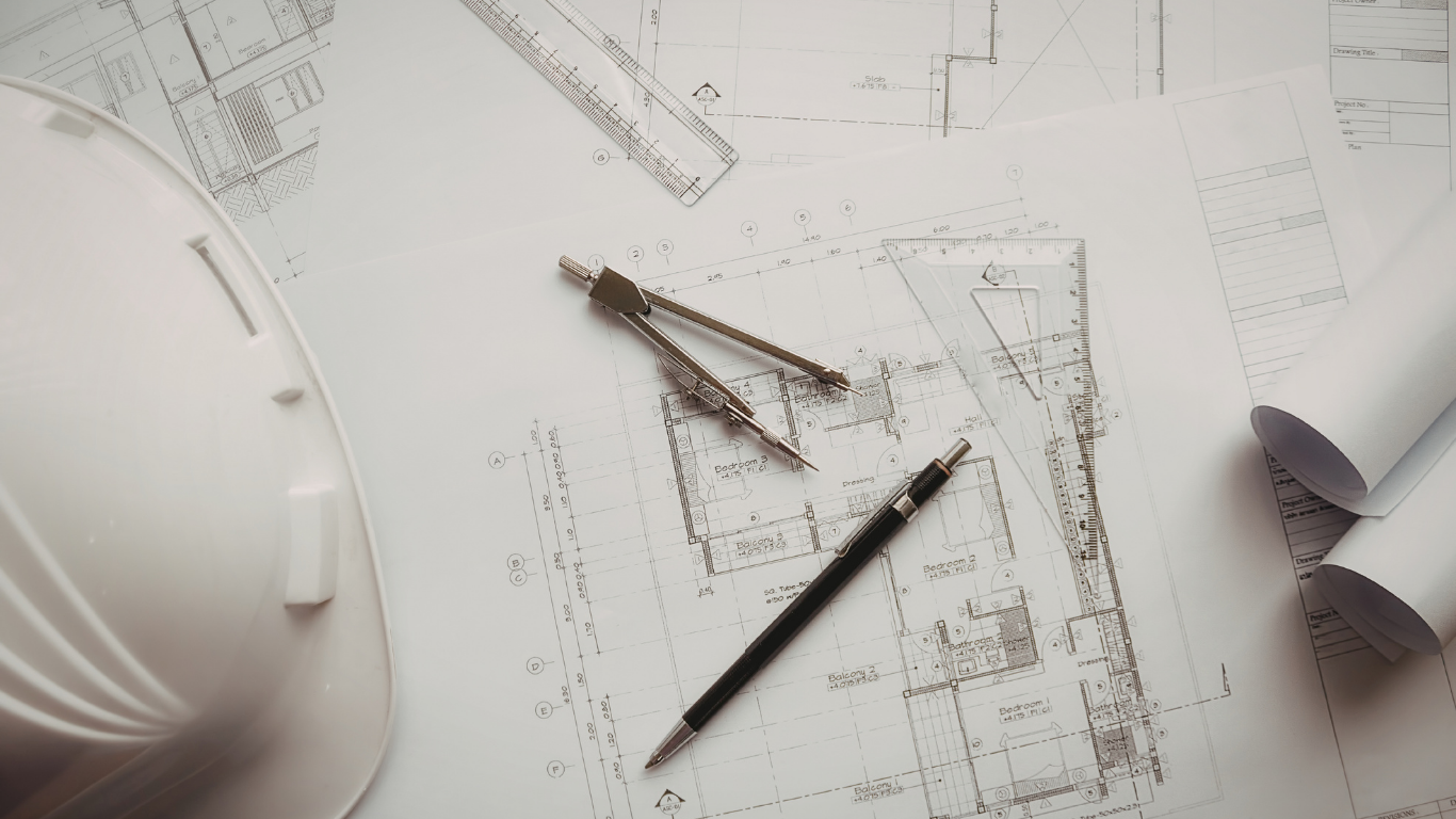 A white hard hat sits beside a technical drafting compass, a pencil, and rolled blueprints on a table.