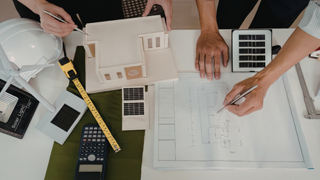 Architects reviewing blueprints and a building model on a table with a calculator, tape measure, and solar panel mockup.