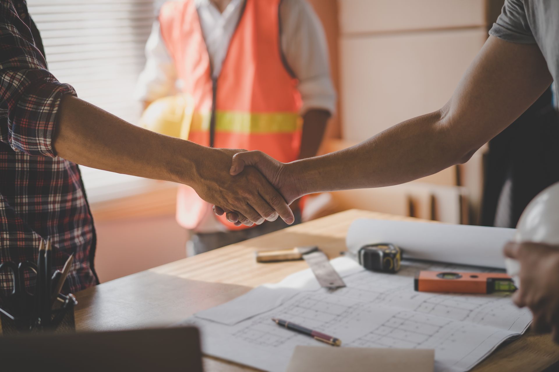 Two people shaking hands over construction plans on a table with tools.