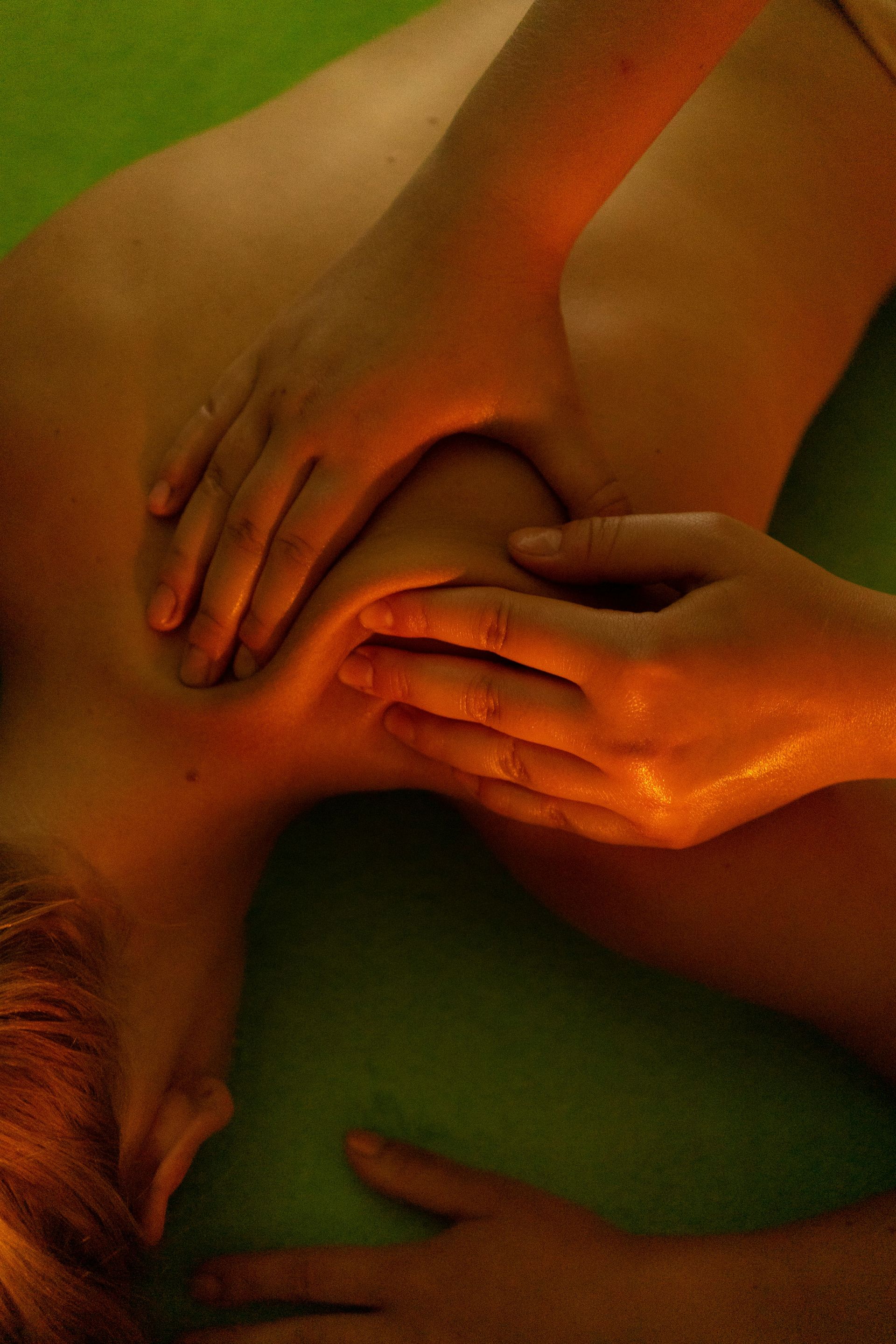 Hands massaging a person's shoulder and neck on a green surface, lit with warm tones.