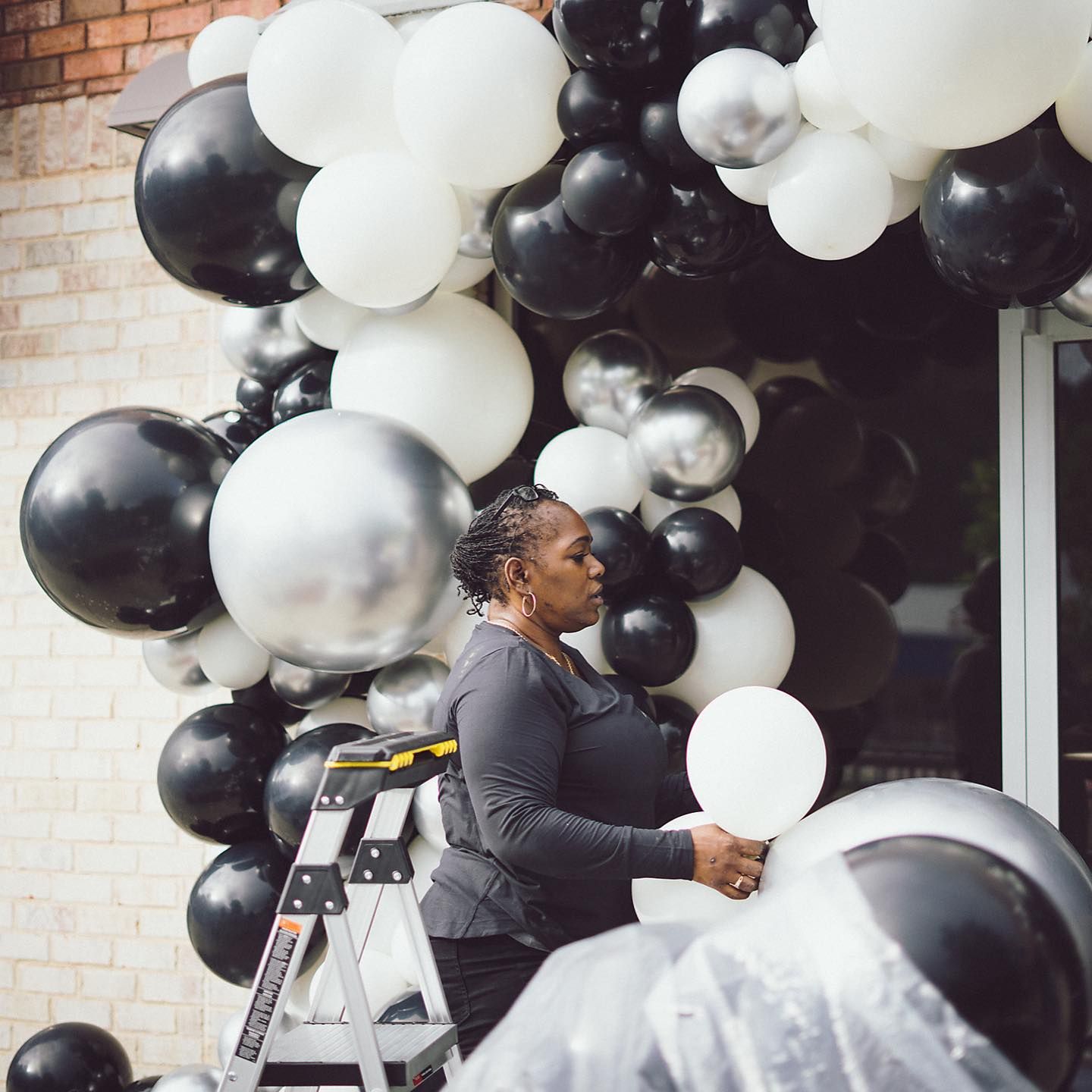 Woman on ladder arranging black, white, and silver balloons around a doorway.