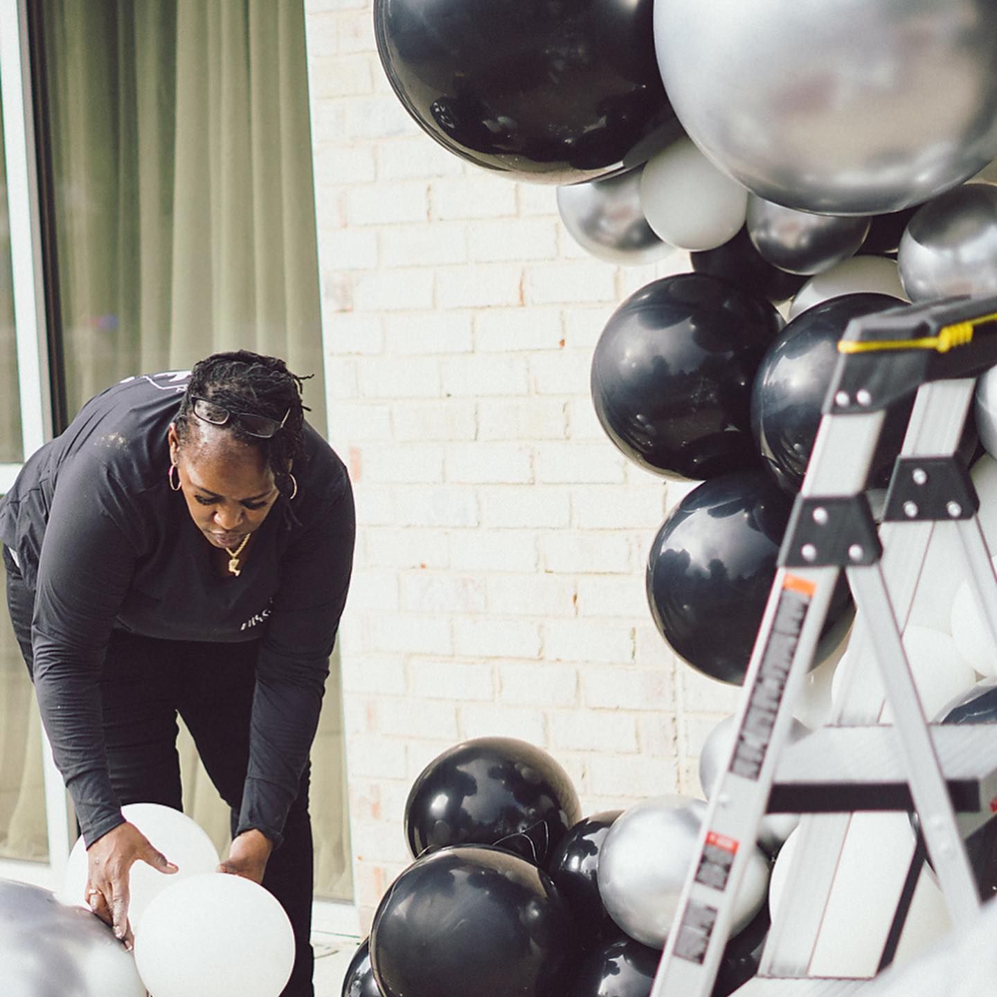 Woman arranging white balloons with black and silver balloons, next to a ladder.