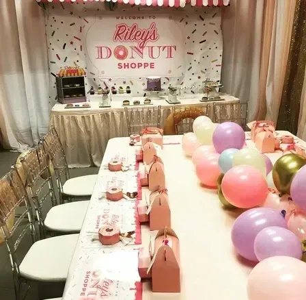Donut-themed party setup with table, chairs, decorations, balloons, and Riley's Donut Shoppe sign.