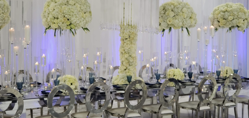 Elegant wedding reception with floral arrangements, candles, and mirrored table. Silver chairs and blue accents.