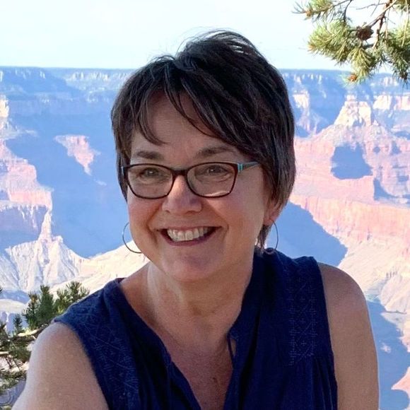 A woman wearing glasses and a blue shirt is smiling in front of a grand canyon.