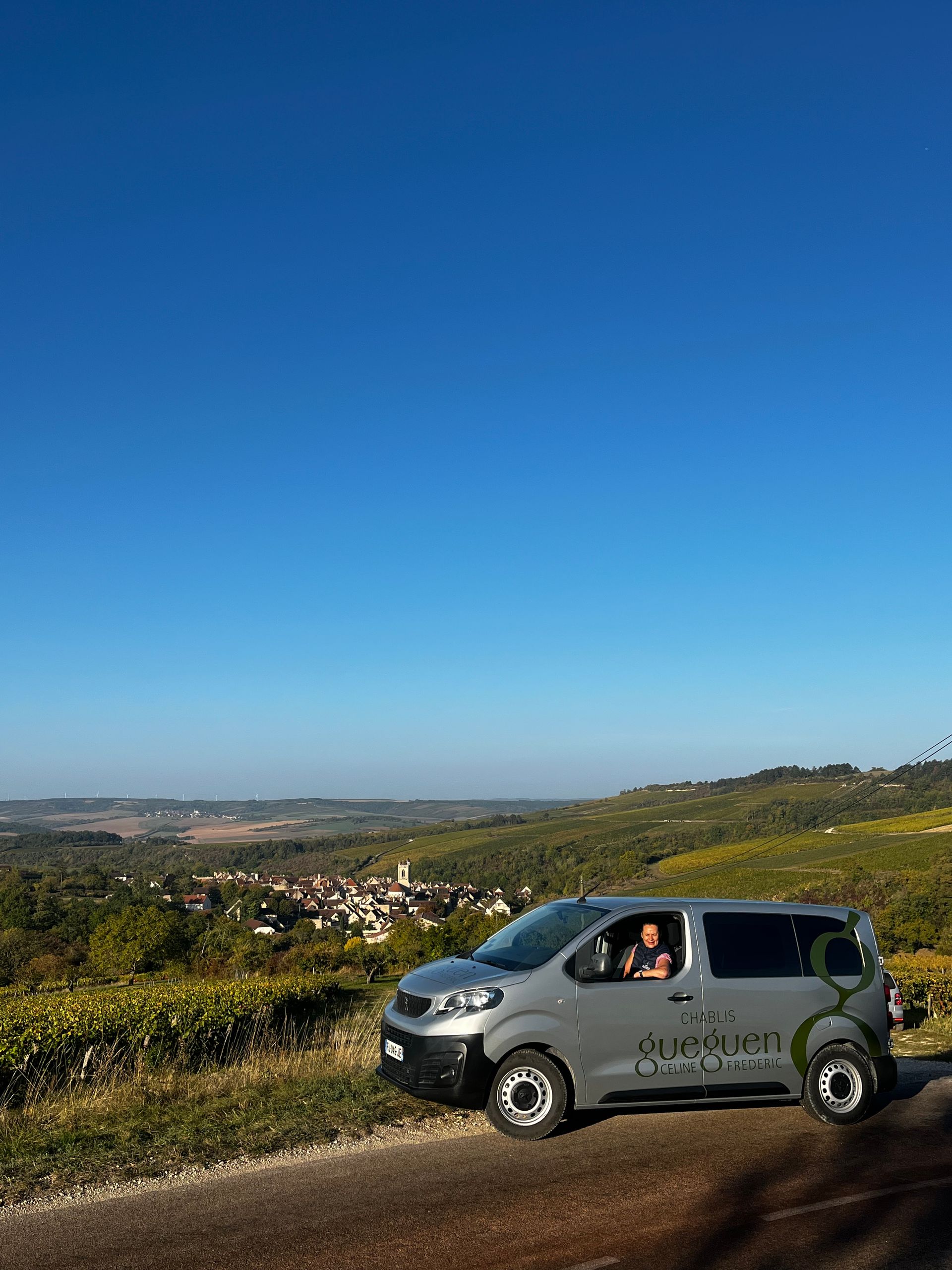 Une camionnette argentée garée sur une route à flanc de colline surplombant une vallée de vignobles et un petit village, sous un ciel d'un bleu limpide.
