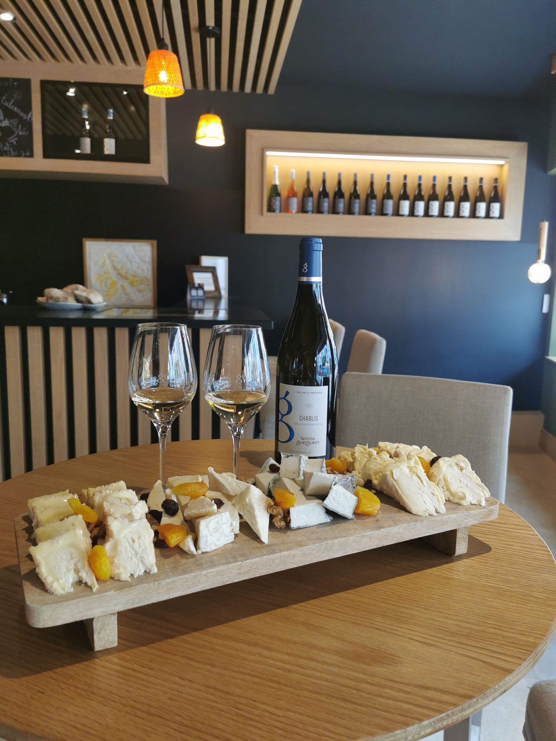 Sur une table ronde, une planche en bois garnie d'un assortiment de fromages et de fruits secs, à côté de deux verres à vin et d'une bouteille.