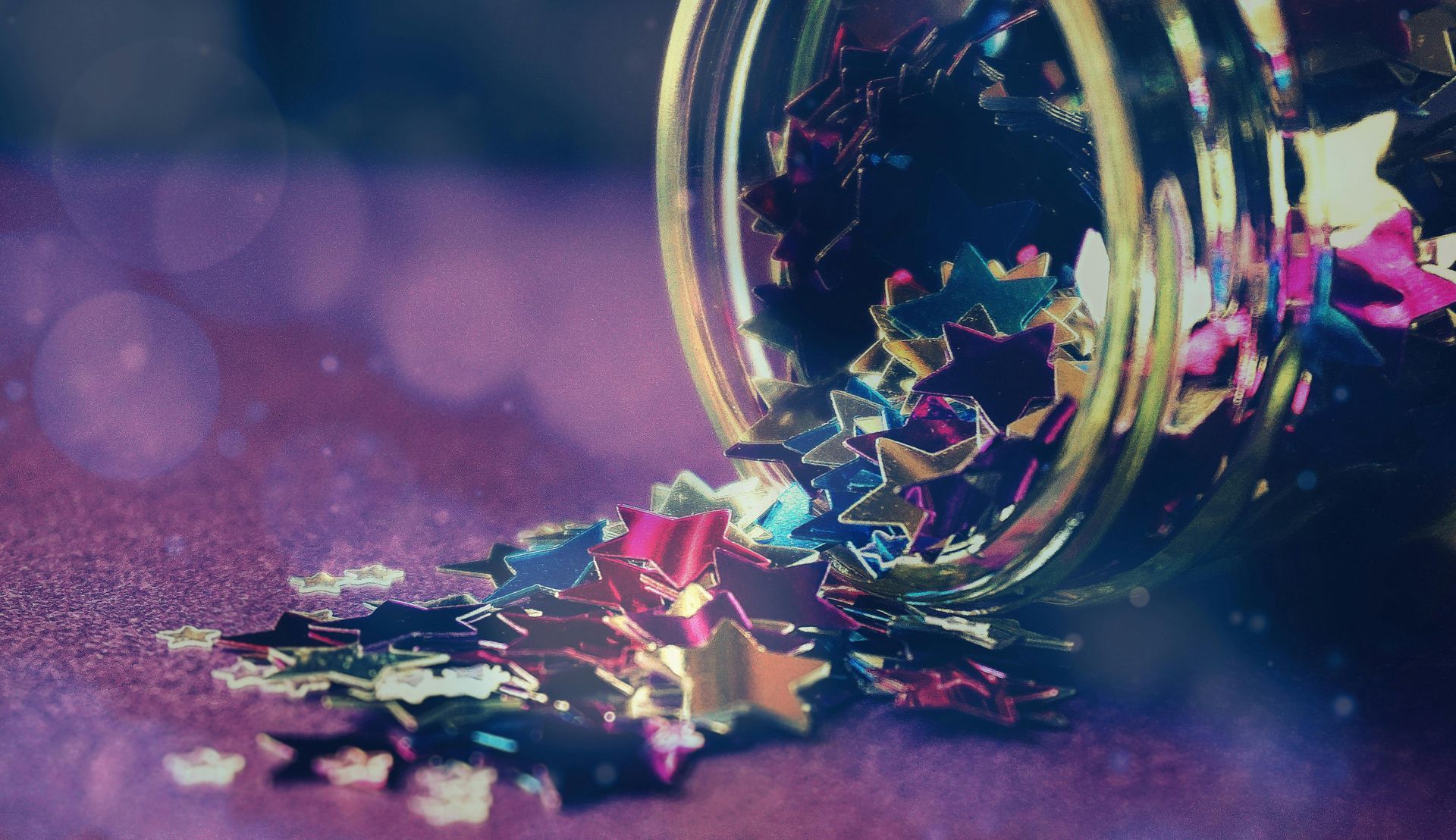 Jar of star-shaped confetti spilling onto a sparkly purple surface; the jar is tilted.