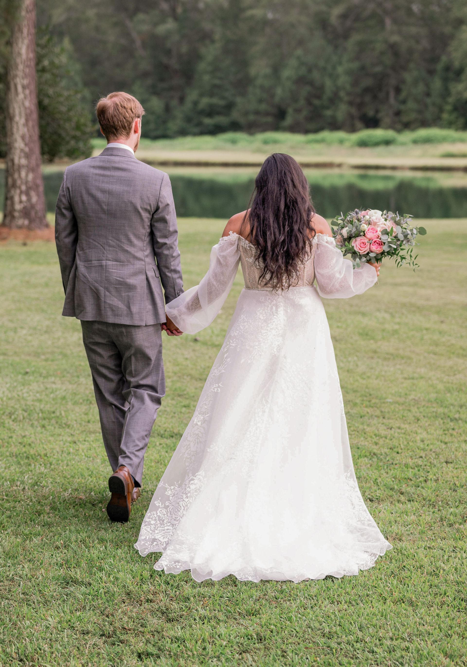 A bride and groom are walking through a grassy field holding hands.