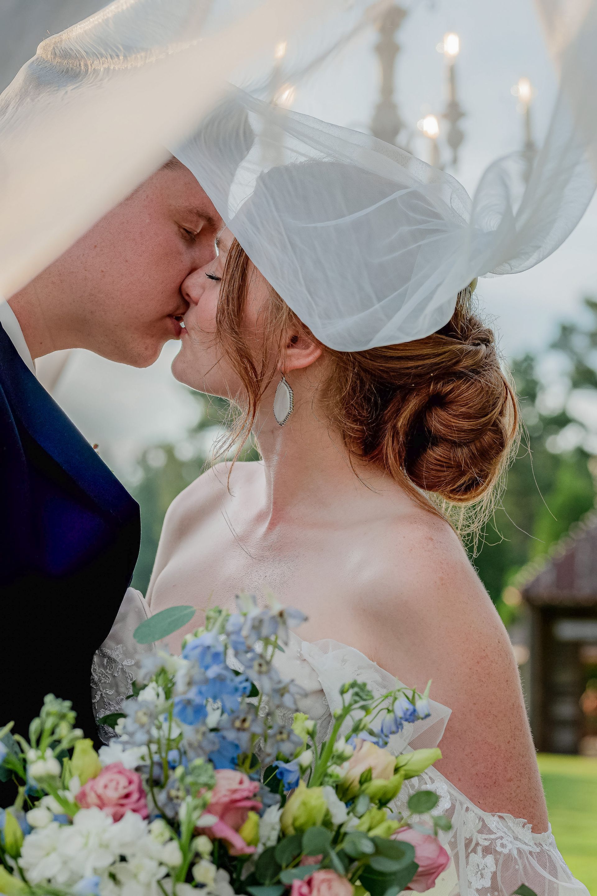 A bride and groom kissing under a chandelier while holding a bouquet of flowers.