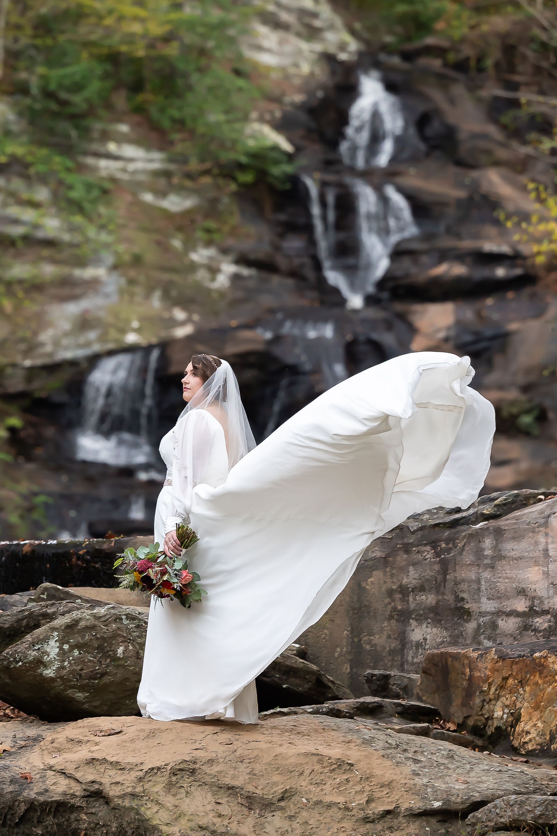 A bride in a wedding dress is standing in front of a waterfall.