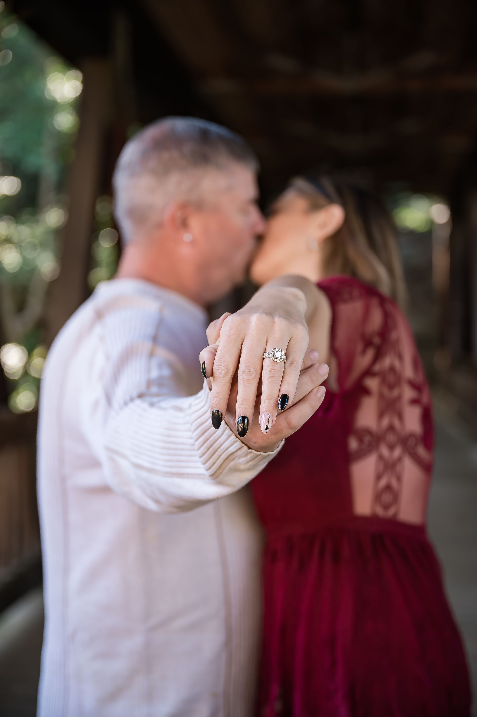 A man and woman are kissing under a bridge while the woman shows off her engagement ring.