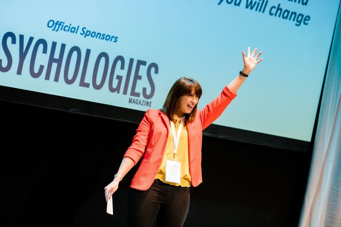 Woman on stage giving a presentation, wearing a coral blazer before a SYCHOLOGIES backdrop