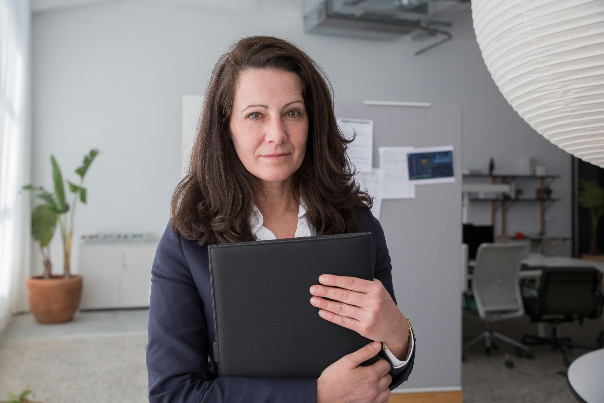 Woman holding a black clipboard in a modern office, with a potted plant and workstations in the background.
