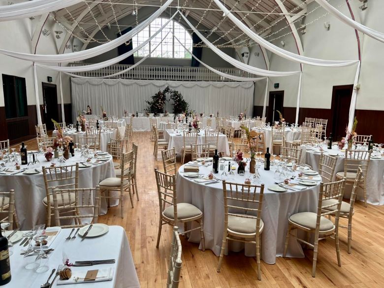 Elegant banquet hall with round white-clothed tables, gold chairs, and draped ceiling decor آماده