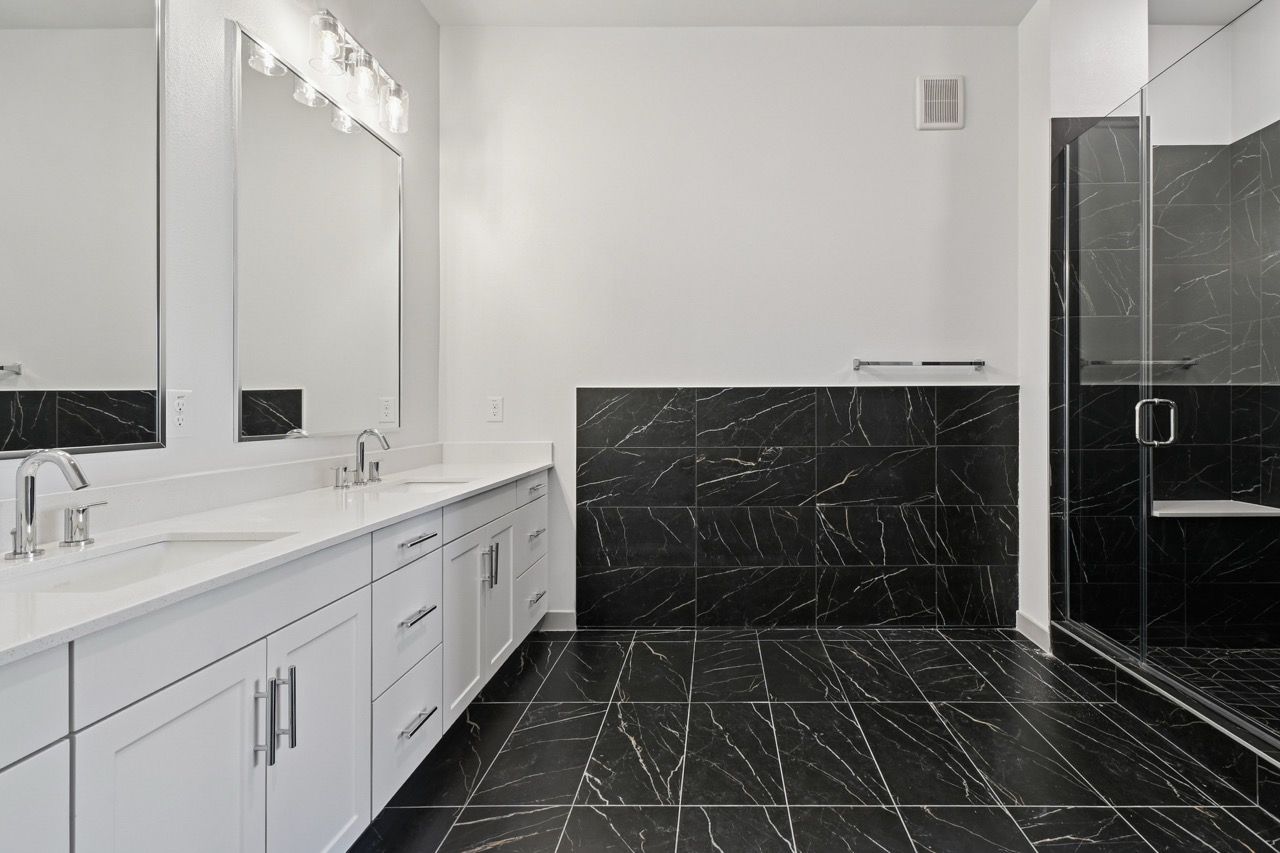Bright bathroom with double vanity, white cabinets, large mirror, and glass shower with black marble tiles  at Ennova Fulshear in Fulshear, TX.