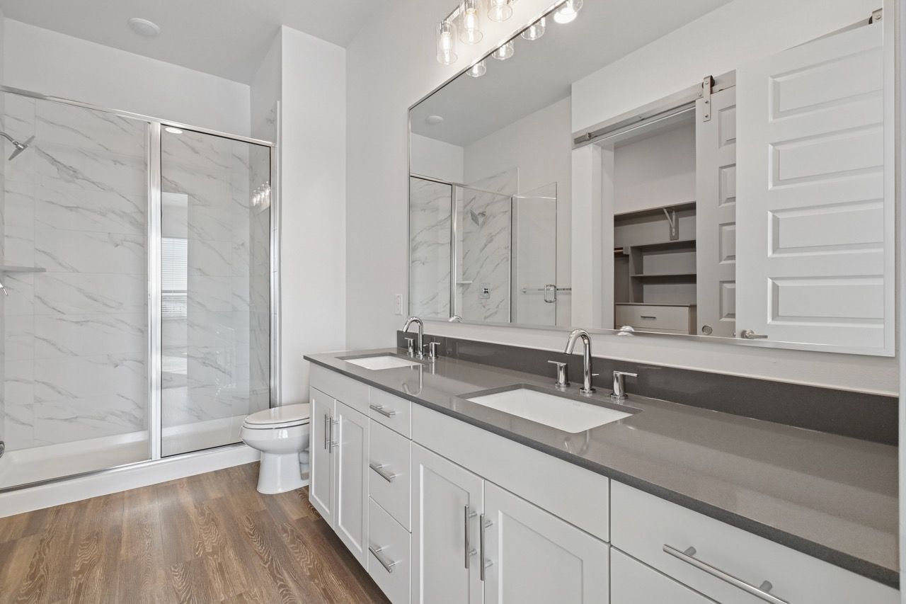 Bathroom with double vanity, gray countertop, glass shower, and marble-tile walls  at Ennova Fulshear in Fulshear, TX.