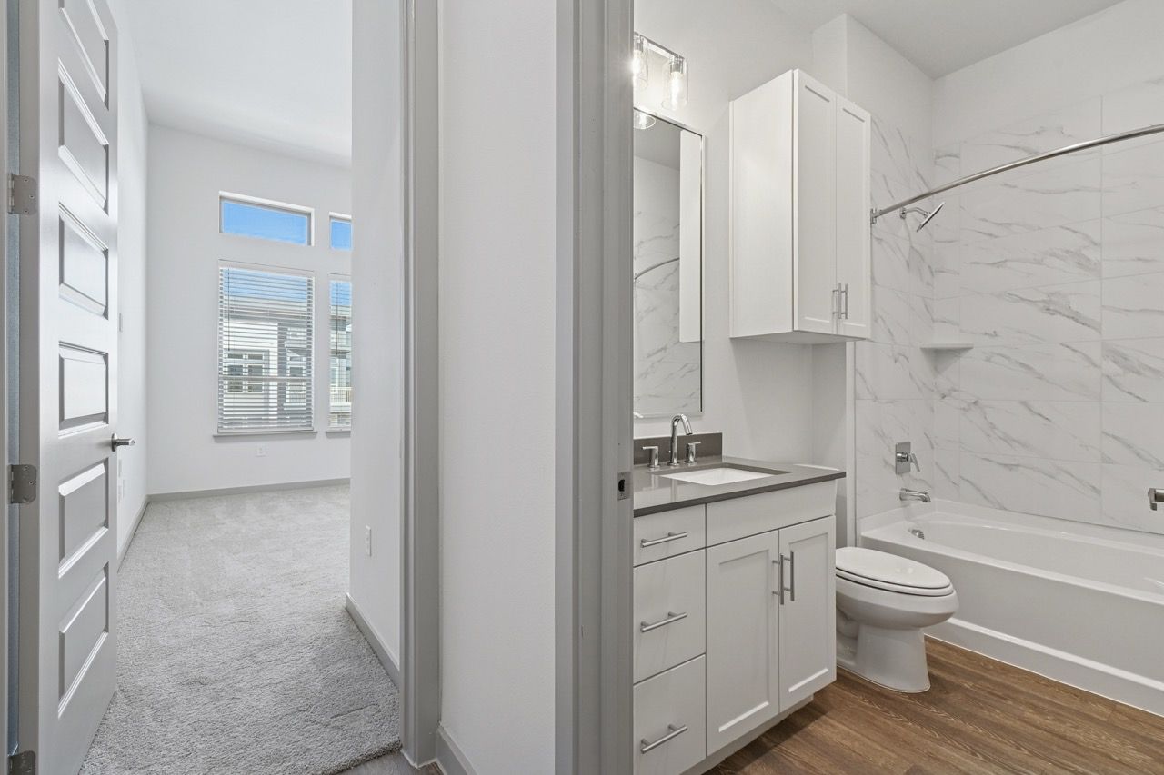 Modern apartment bathroom with marble-tile surround, white vanity, and adjacent carpeted living space  at Ennova Fulshear in Fulshear, TX.