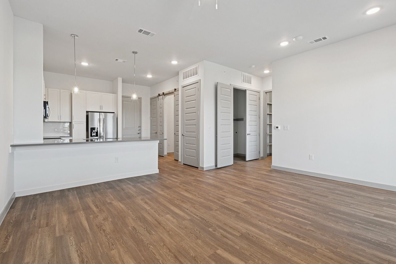 Open-plan kitchen and living area with white cabinets, stainless steel fridge, and wood-look flooring  at Ennova Fulshear in Fulshear, TX.