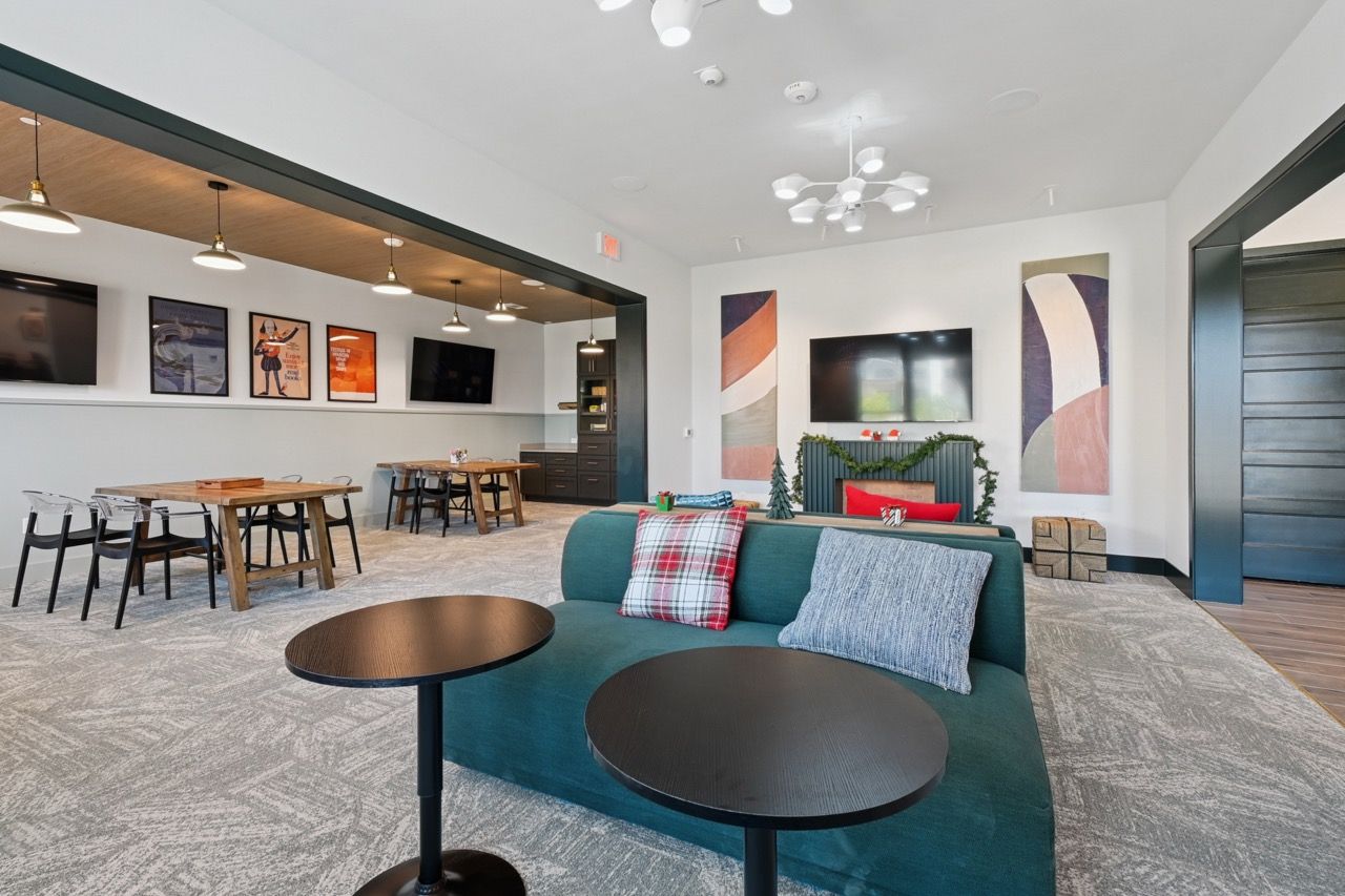 Open community lounge with teal sofa, round coffee tables, wall art, and wall-mounted TVs  at Ennova Fulshear in Fulshear, TX.