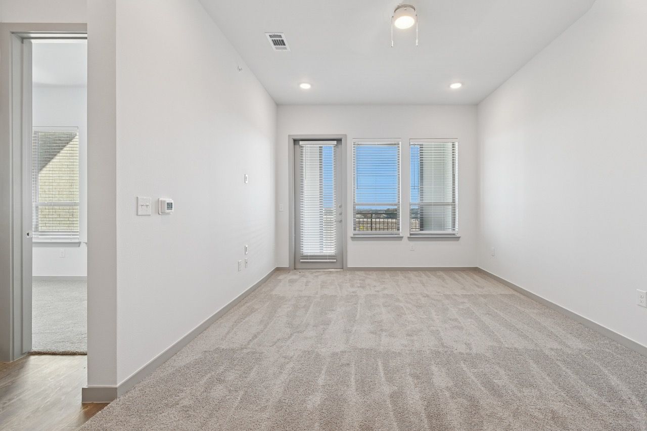 Unfurnished living room with white walls, beige carpet, blinds, and a balcony door  at Ennova Fulshear in Fulshear, TX.