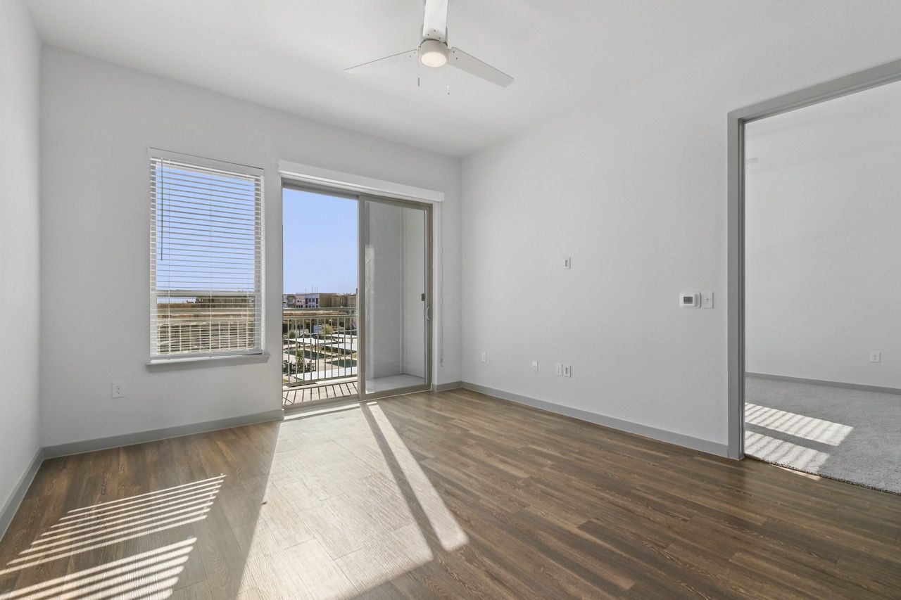 Empty living room with wood-look flooring, ceiling fan, and sliding door to balcony  at Ennova Fulshear in Fulshear, TX.