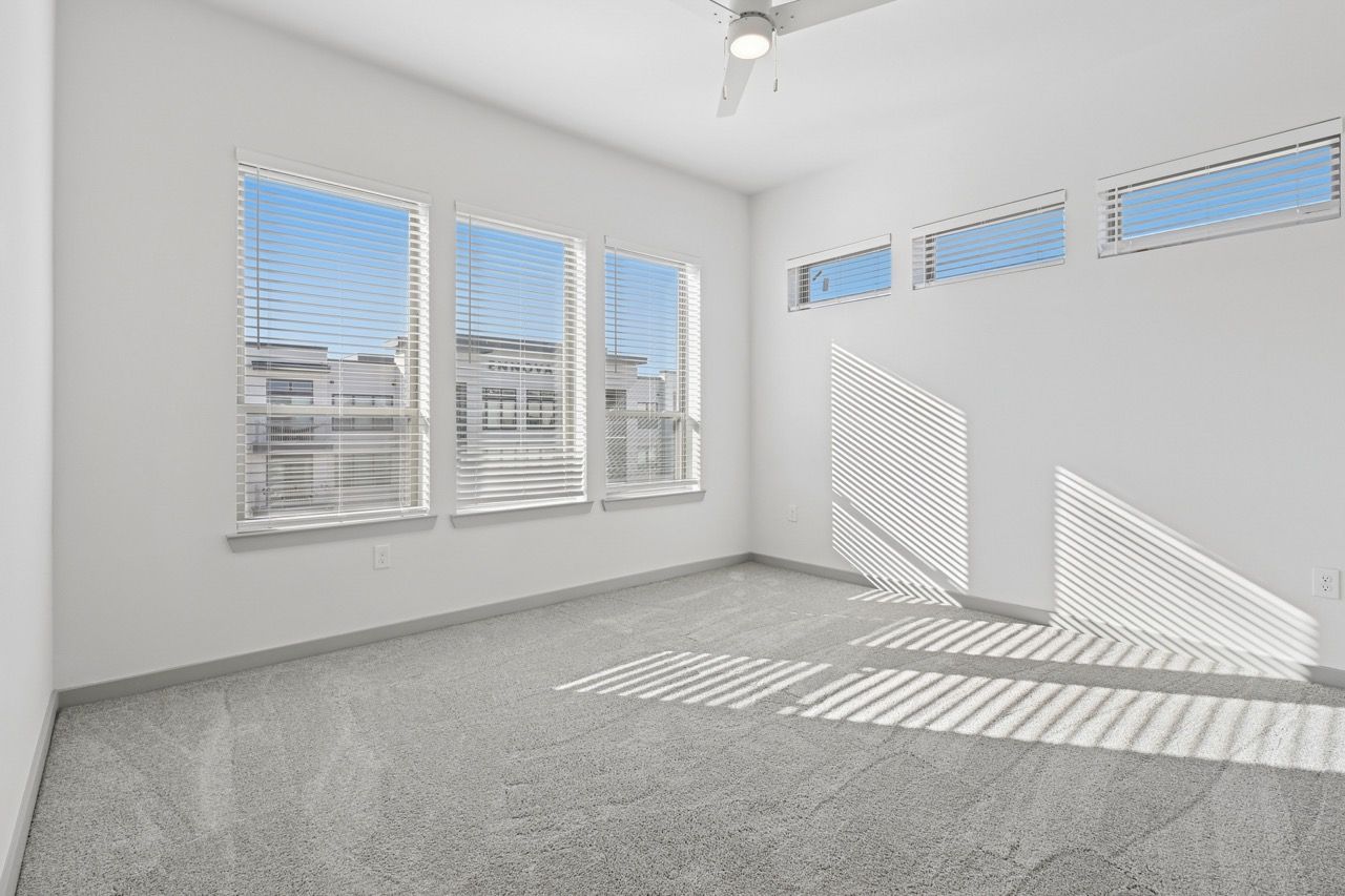 Bright, empty apartment room with white walls, carpeting, ceiling fan, and multiple windows with blinds  at Ennova Fulshear in Fulshear, TX.