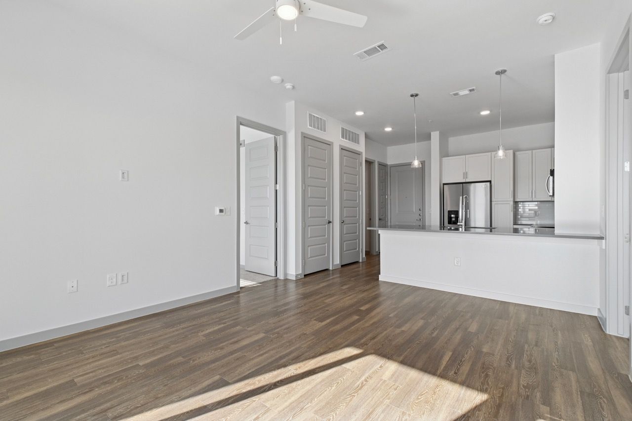 Open living area with hardwood floors, white walls, and a modern kitchen with an island  at Ennova Fulshear in Fulshear, TX.