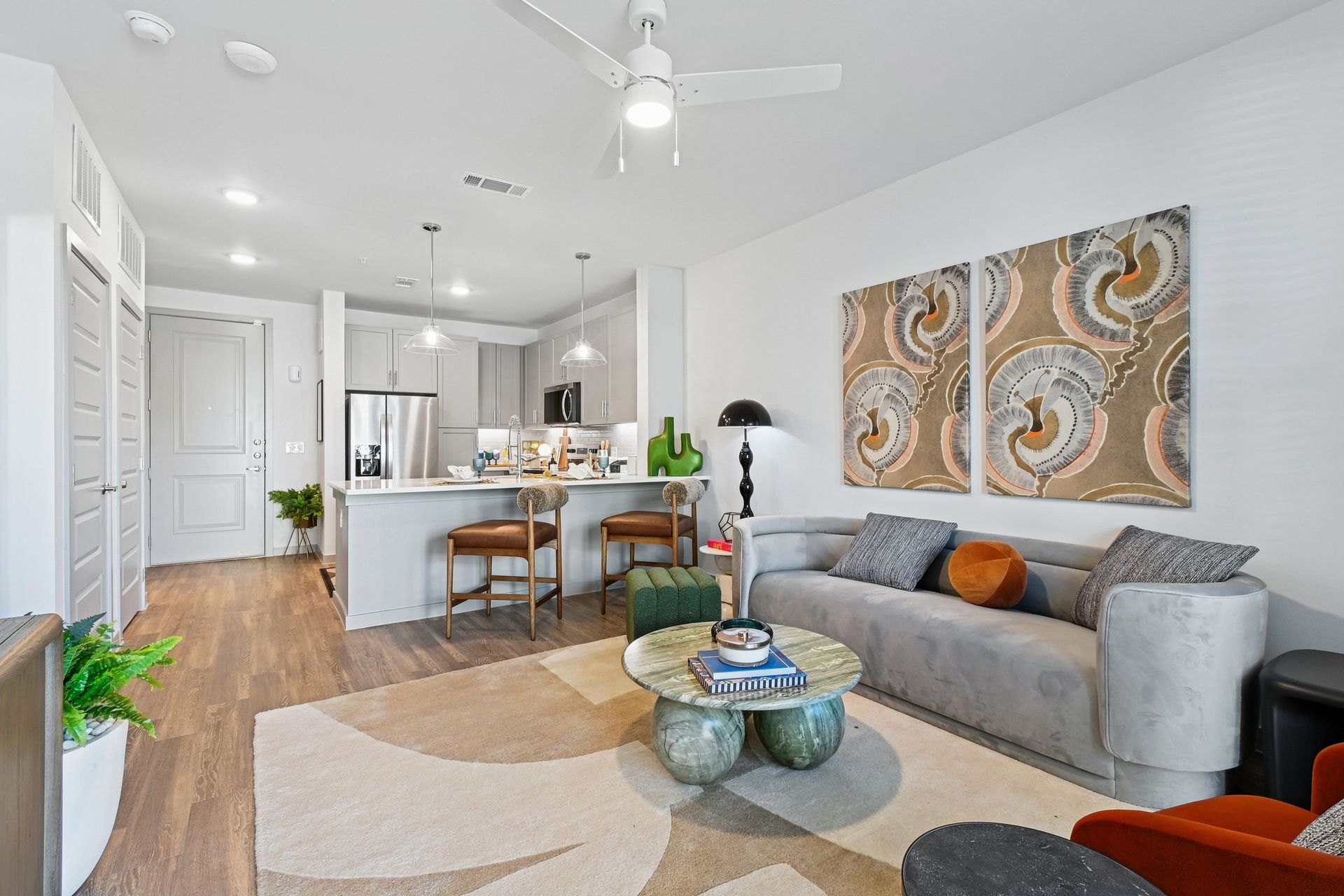 Modern living room with kitchen in the background. Light gray and brown furniture, artwork, and a patterned rug.