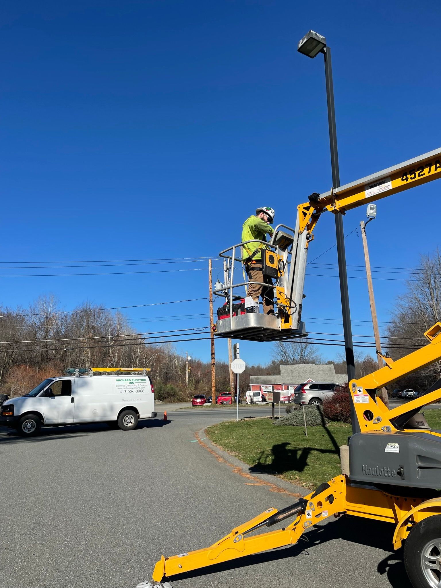 Worker in lift bucket repairing a streetlight under a blue sky, white van nearby.