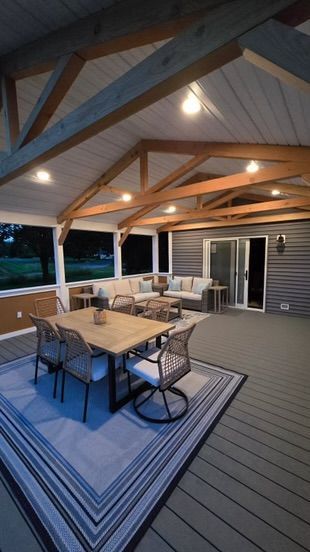 Screened-in porch with dining table and seating area, wooden beams and light fixtures, grey siding, and blue rug.