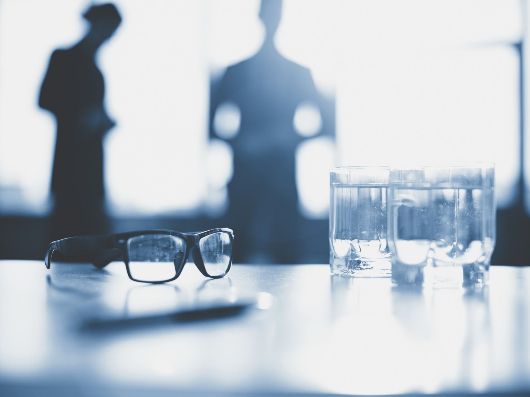 Glasses, pen, and two water glasses on a table; blurred figures in background.