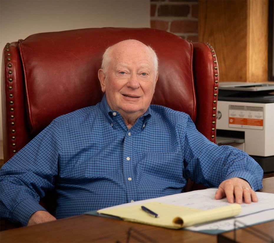 An older man is sitting in a red leather chair at a desk.