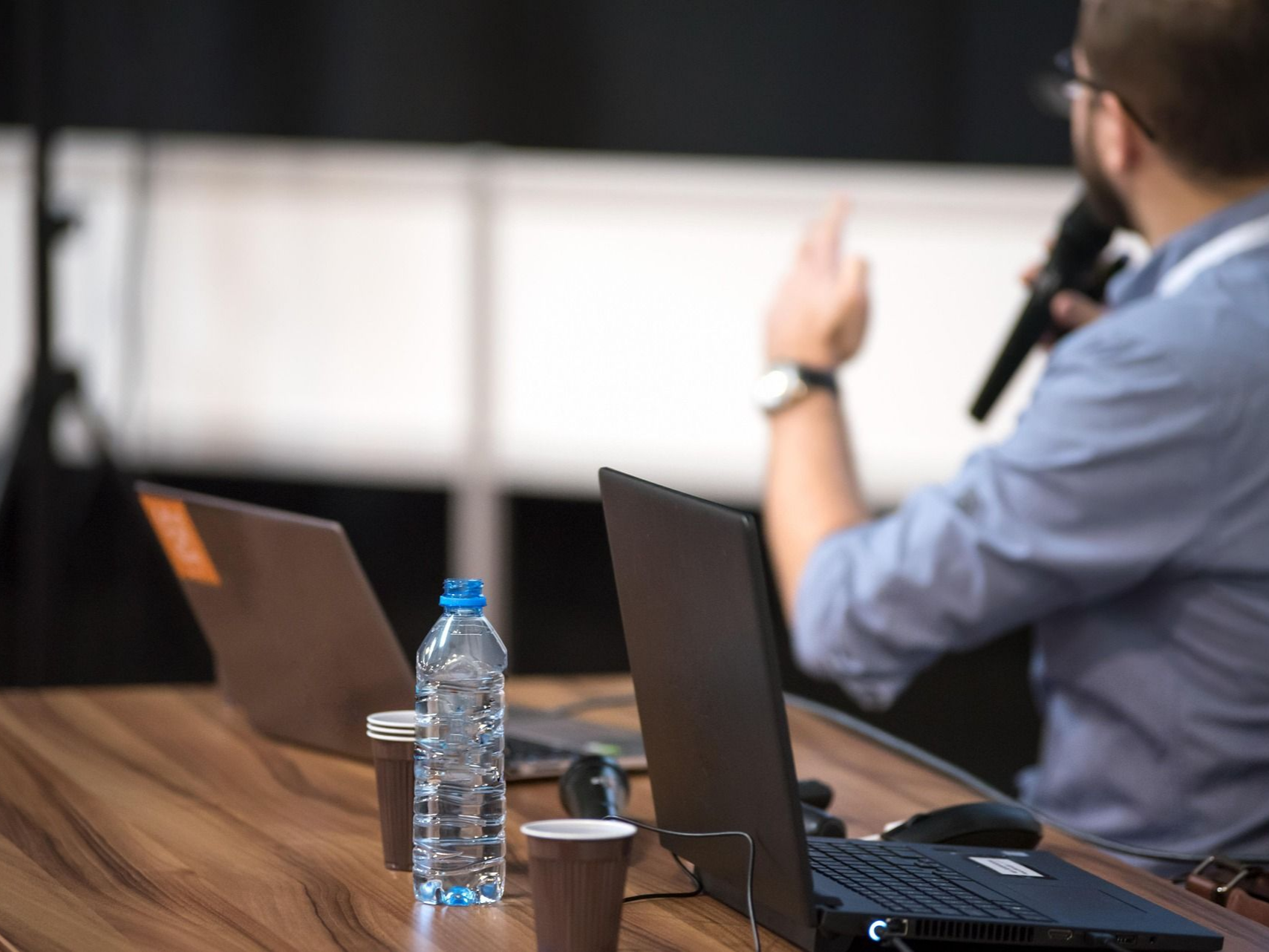 Man speaking at event, holding microphone. Laptops, water bottle on table in front. White screen backdrop.