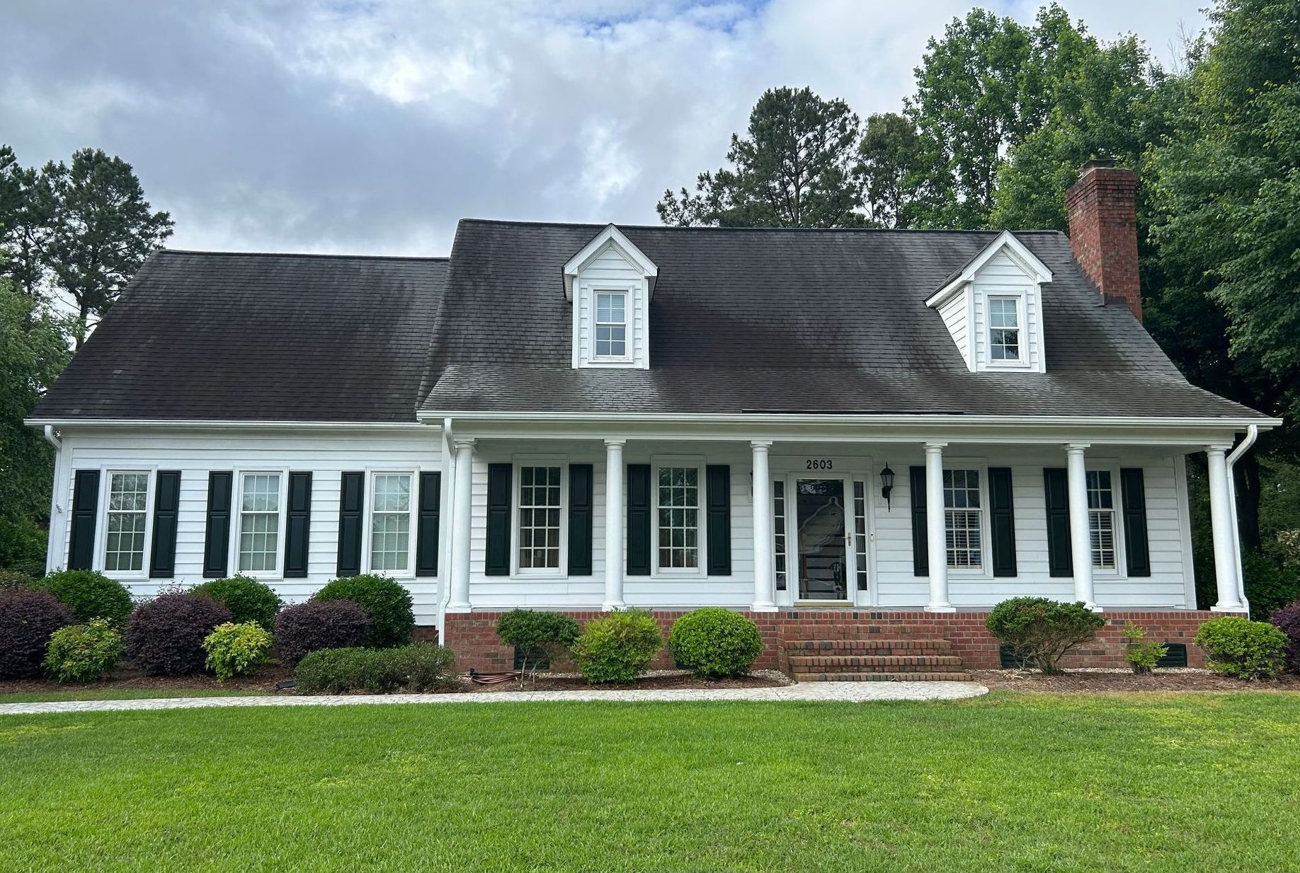 White house with black shutters, red brick chimney, and green lawn under cloudy skies.