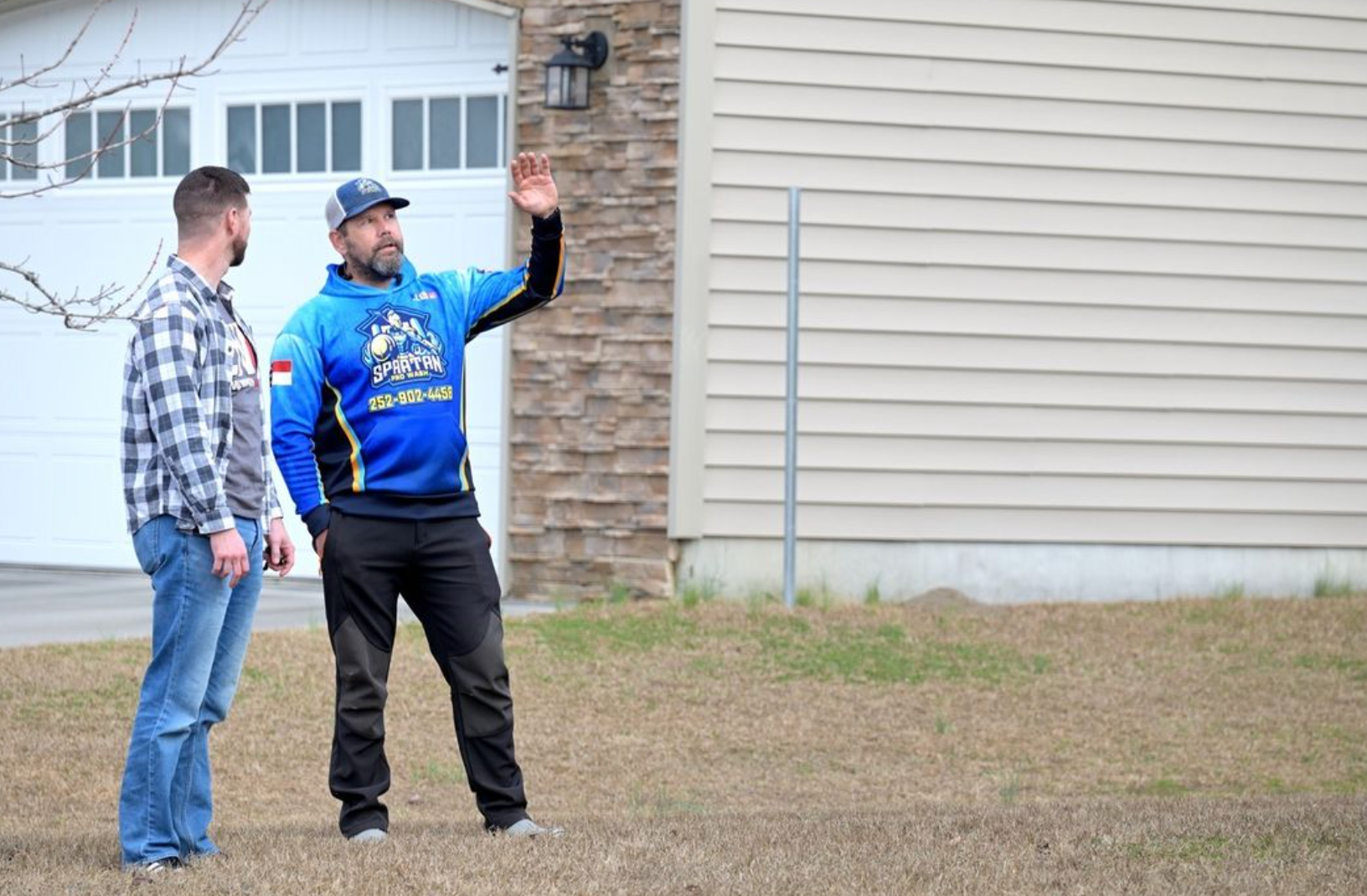 Two men standing outside a beige house. One gestures with his arm up.