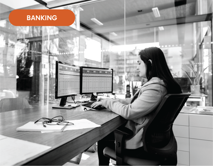 A woman is sitting at a desk in front of two computer monitors.