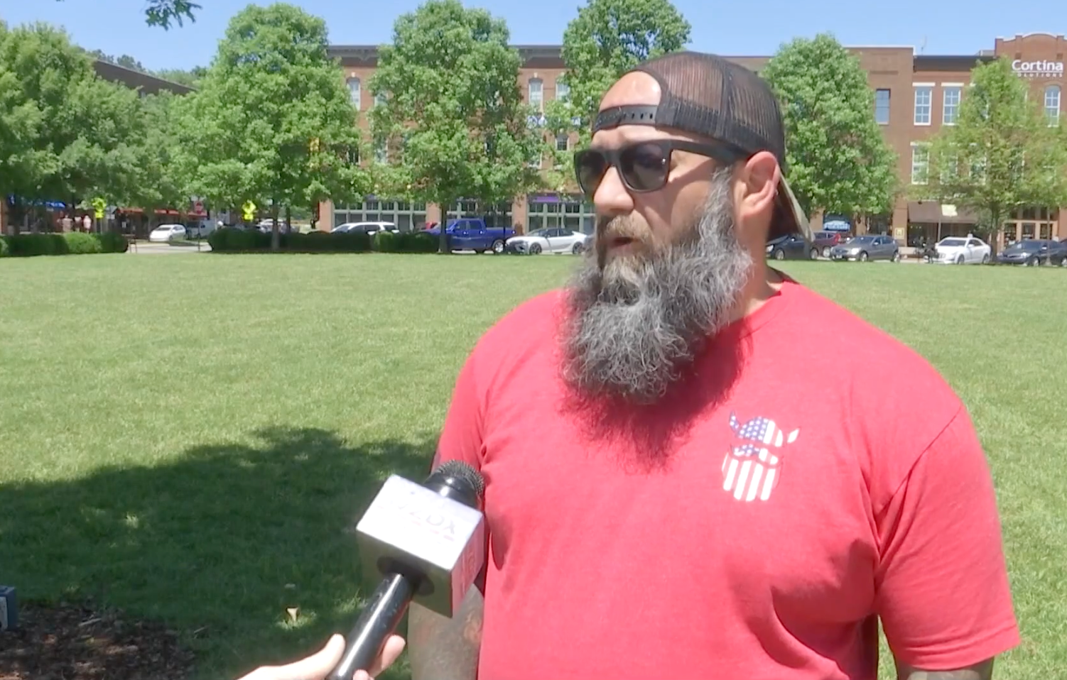 A man with a beard is talking into a microphone in a park.